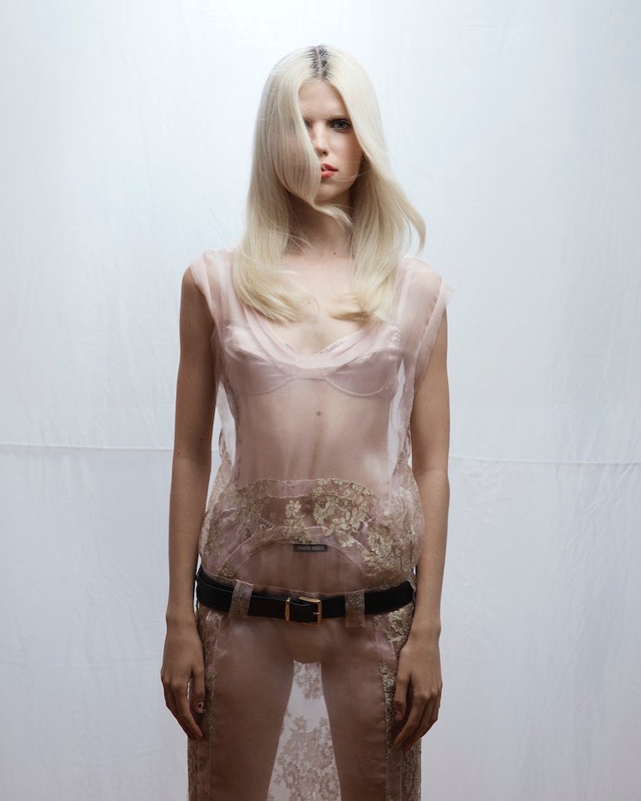 Blonde woman wearing a sheer pale pink dress with a black belt against a plain white seamless studio background.