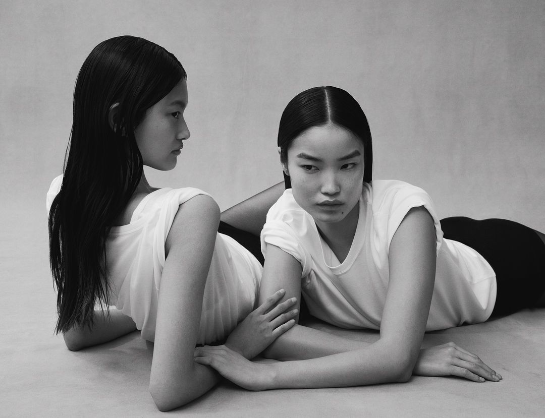 Two Asian women with black hair and white t-shirts lie on a light gray surface, their hands touching, in a fashion editorial studio shot.