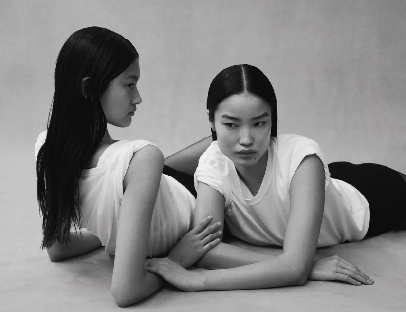Two Asian women with black hair and white t-shirts lie on a light gray surface, their hands touching, in a fashion editorial studio shot.