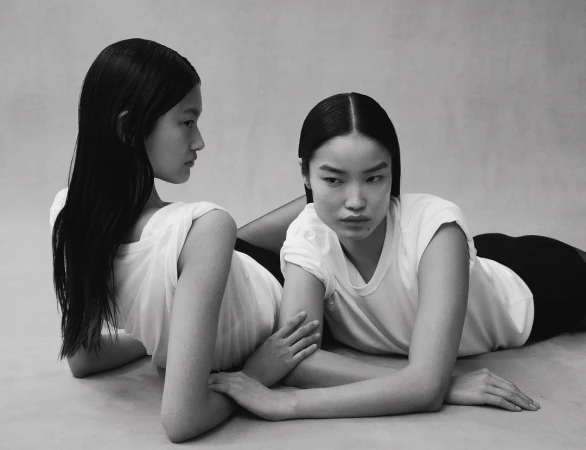 Two Asian women with black hair and white t-shirts lie on a light gray surface, their hands touching, in a fashion editorial studio shot.