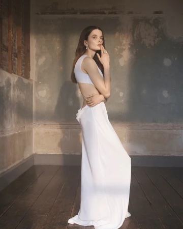 Elegant woman in white dress poses in naturally lit corner room with distressed walls and dark wood floors, creating a serene ambiance.