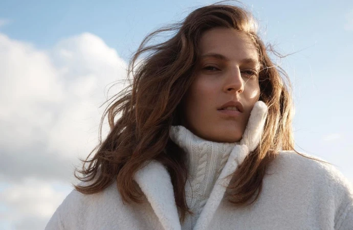 Close-up portrait of a woman with tousled brown hair wearing a white chunky turtleneck sweater against a clear sky background
