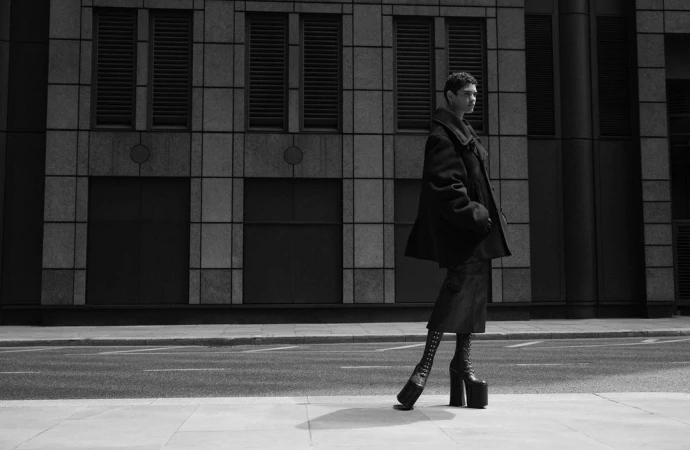 Model in dark oversized coat and platform boots standing on a city street against a stone building facade in monochrome.
