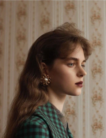 Profile view of a young woman with long wavy brown hair and large floral earrings, wearing a green plaid jacket against a floral wallpaper.