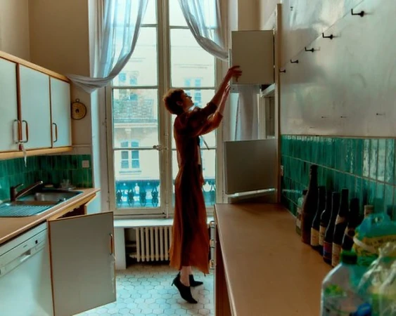 Woman in an orange dress reaching into a wall-mounted cabinet in a kitchen with green tiled walls, photographed by Adrian Mesko