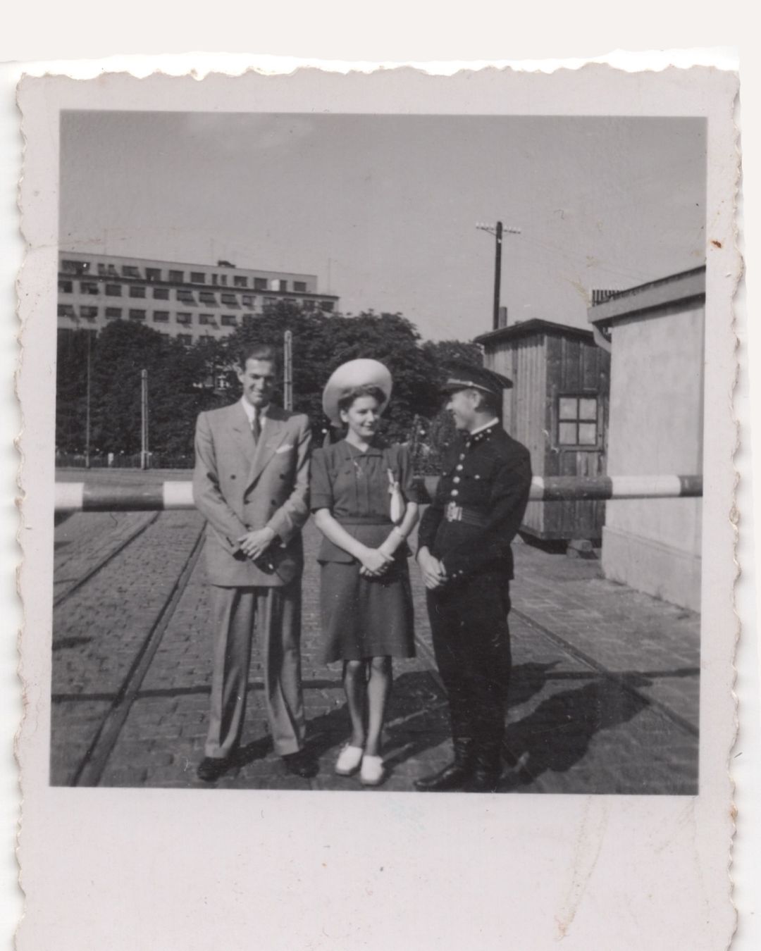Vintage photo of a couple standing with a uniformed guard in front of a gate, with an urban landscape and sky in the background.