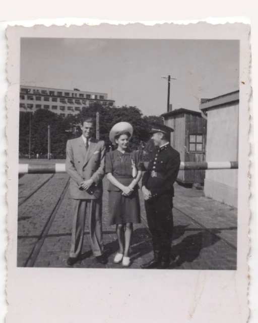 Vintage photo of a couple standing with a uniformed guard in front of a gate, with an urban landscape and sky in the background.