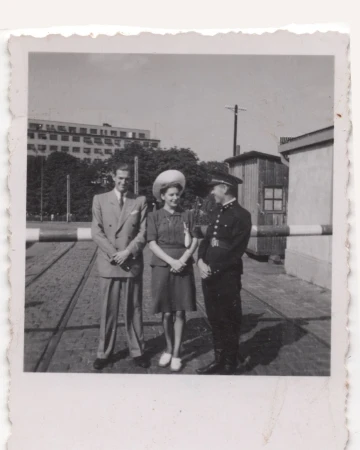 Vintage photo of a couple standing with a uniformed guard in front of a gate, with an urban landscape and sky in the background.