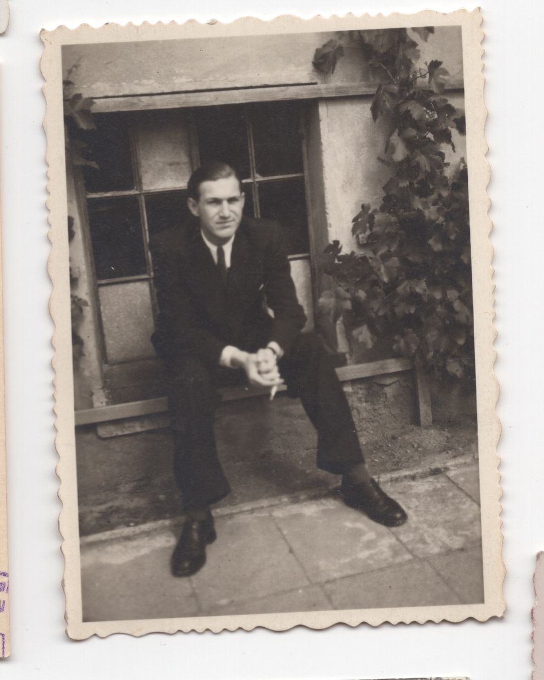 Vintage monochrome photograph of a seated man in a dark suit against an exterior wall with climbing ivy.