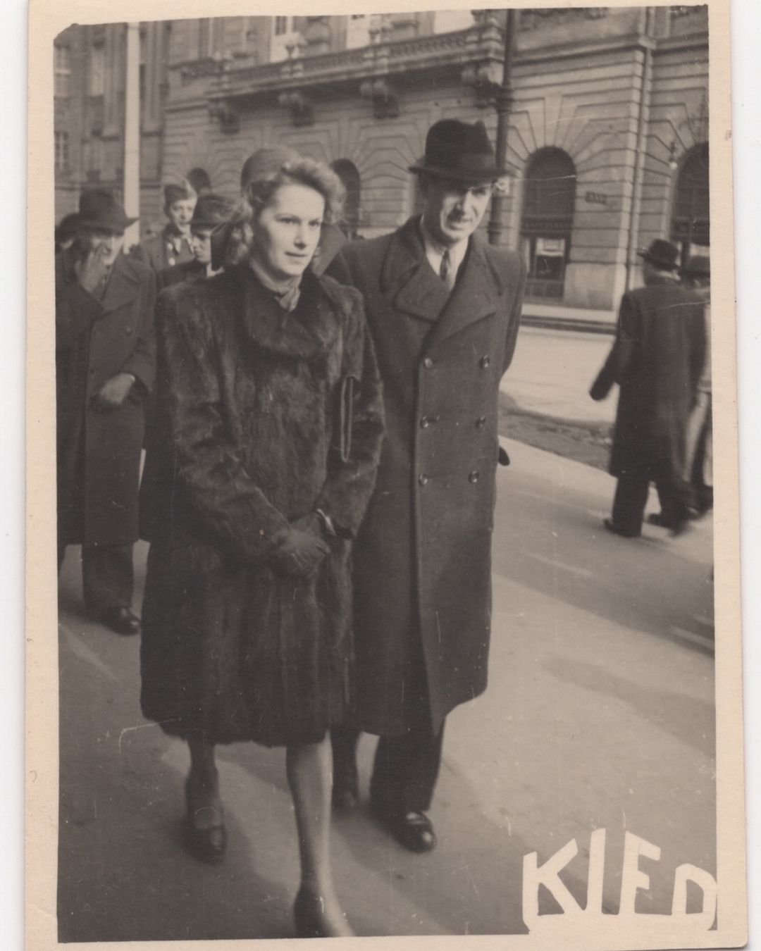 Vintage photo features a well-dressed couple walking on a city street, with other pedestrians and a building in the background.