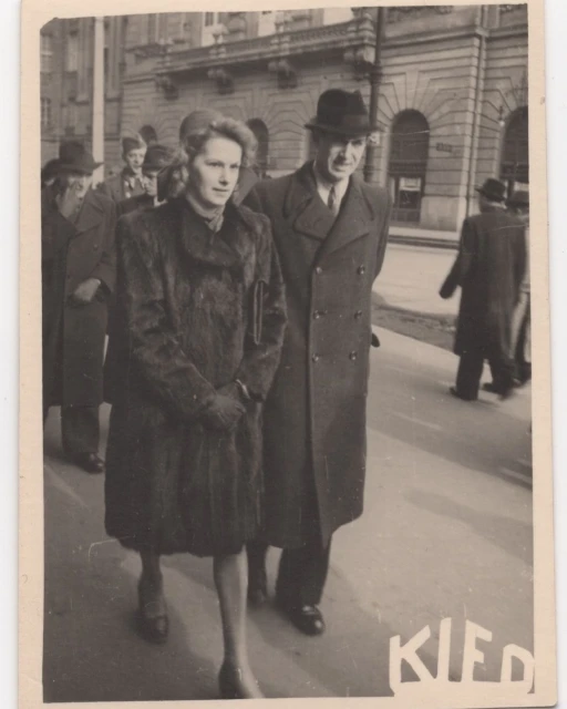 Vintage photo features a well-dressed couple walking on a city street, with other pedestrians and a building in the background.