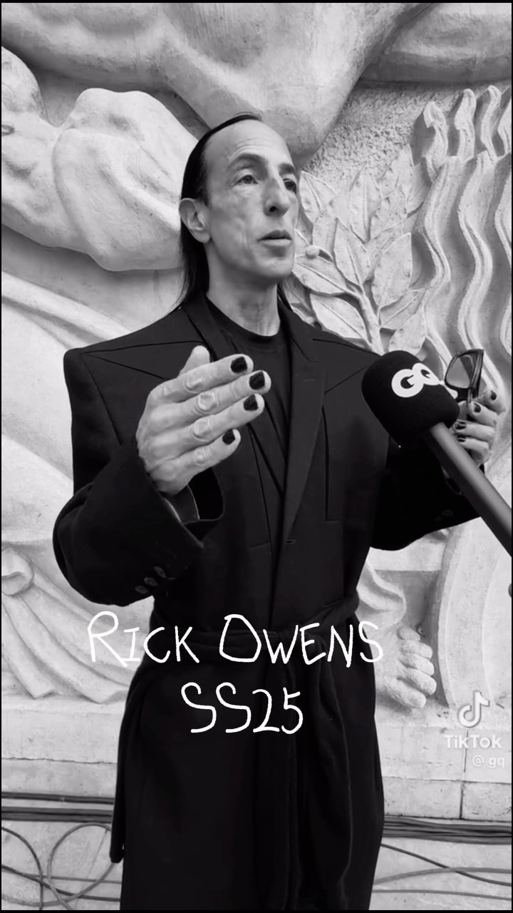 Rick Owens at event in dark suit with microphones, architectural background, manicured nails, black and white image