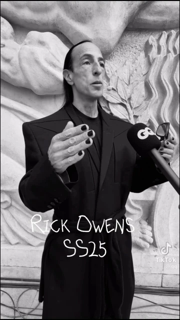 Rick Owens at event in dark suit with microphones, architectural background, manicured nails, black and white image