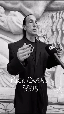 Rick Owens at event in dark suit with microphones, architectural background, manicured nails, black and white image