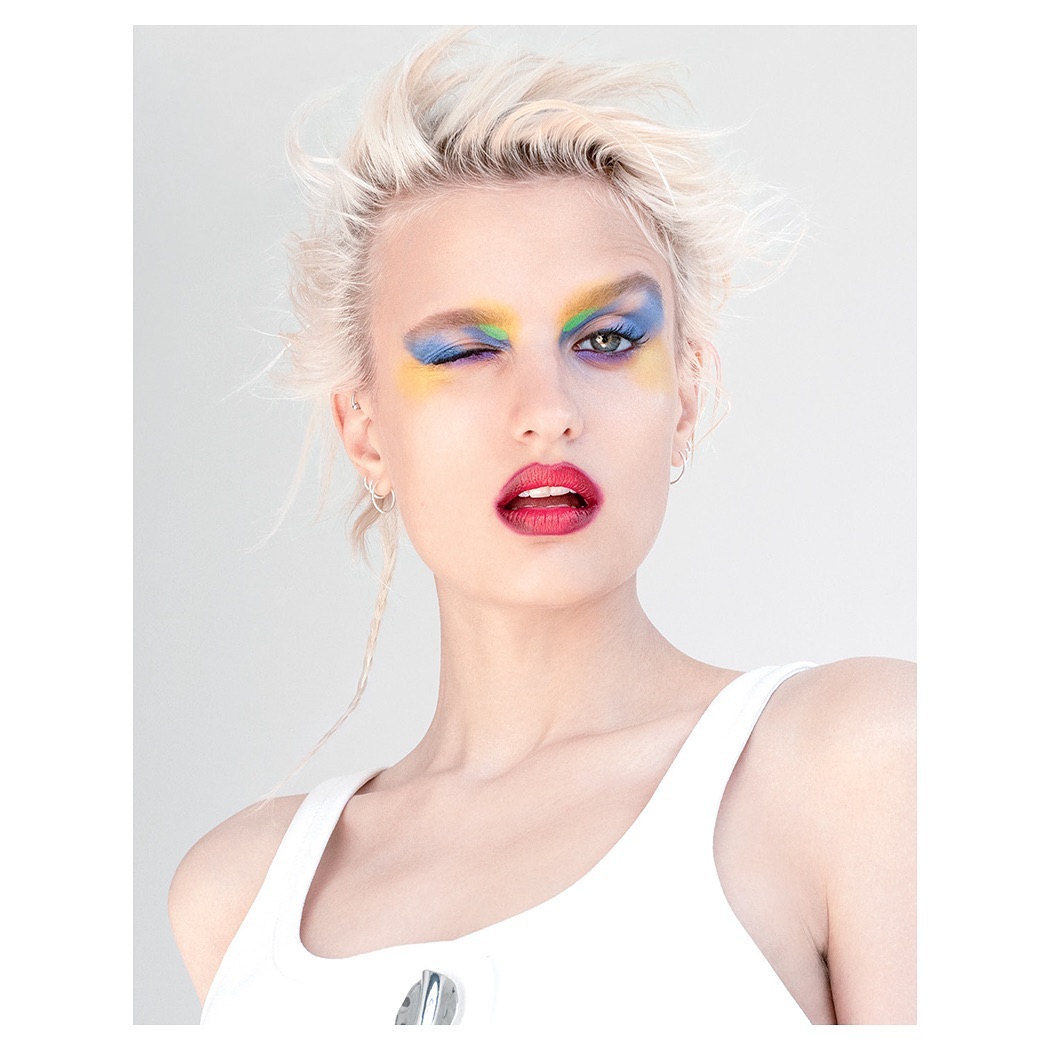 Close-up of a winking woman with blonde hair and colorful eyeshadow in a bright studio setting. Beauty and fashion editorial.