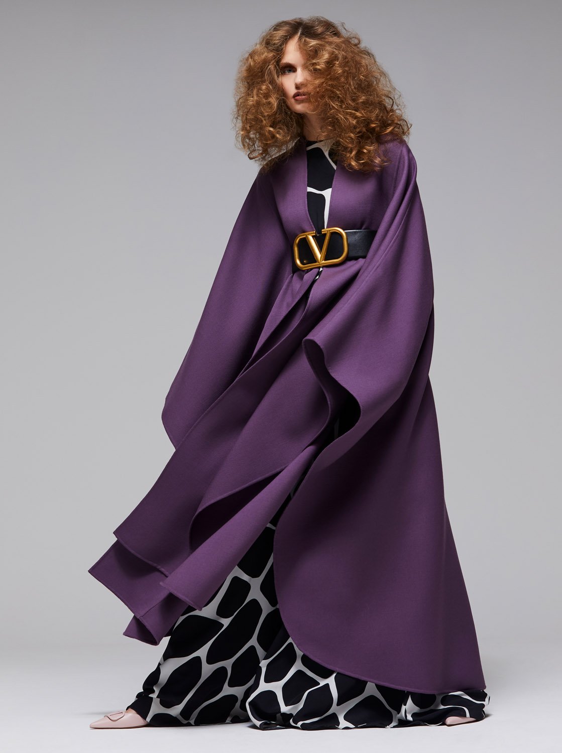 Fashion shot of a woman with curly hair wearing a flowing purple cape with a large gold belt buckle against a grey backdrop.