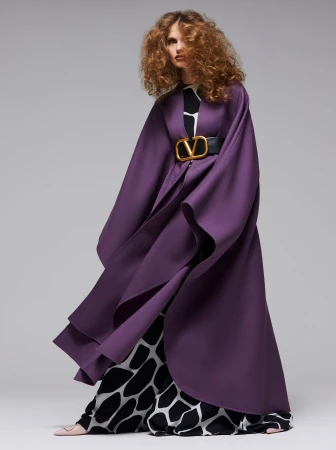 Fashion shot of a woman with curly hair wearing a flowing purple cape with a large gold belt buckle against a grey backdrop.