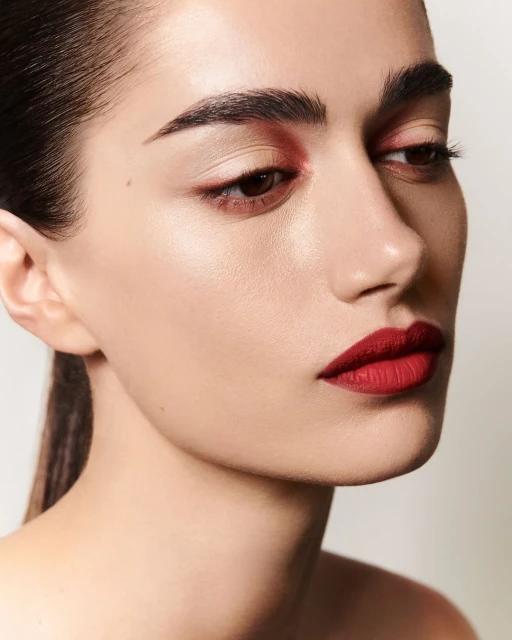 Close-up shot of a woman with red lipstick and red eye shadow, pale skin, slicked back dark hair, facing right in a neutral studio.