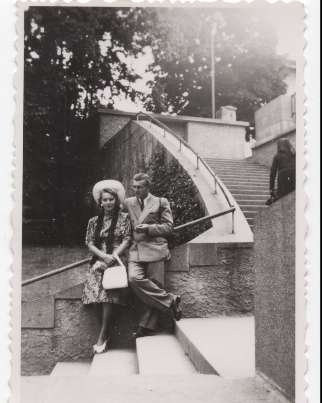 Vintage photo of a stylish couple, an adult male and female, posing outdoors on stone steps near trees and a building.