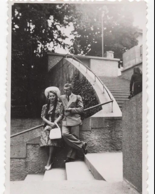 Vintage photo of a stylish couple, an adult male and female, posing outdoors on stone steps near trees and a building.