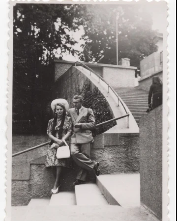Vintage photo of a stylish couple, an adult male and female, posing outdoors on stone steps near trees and a building.