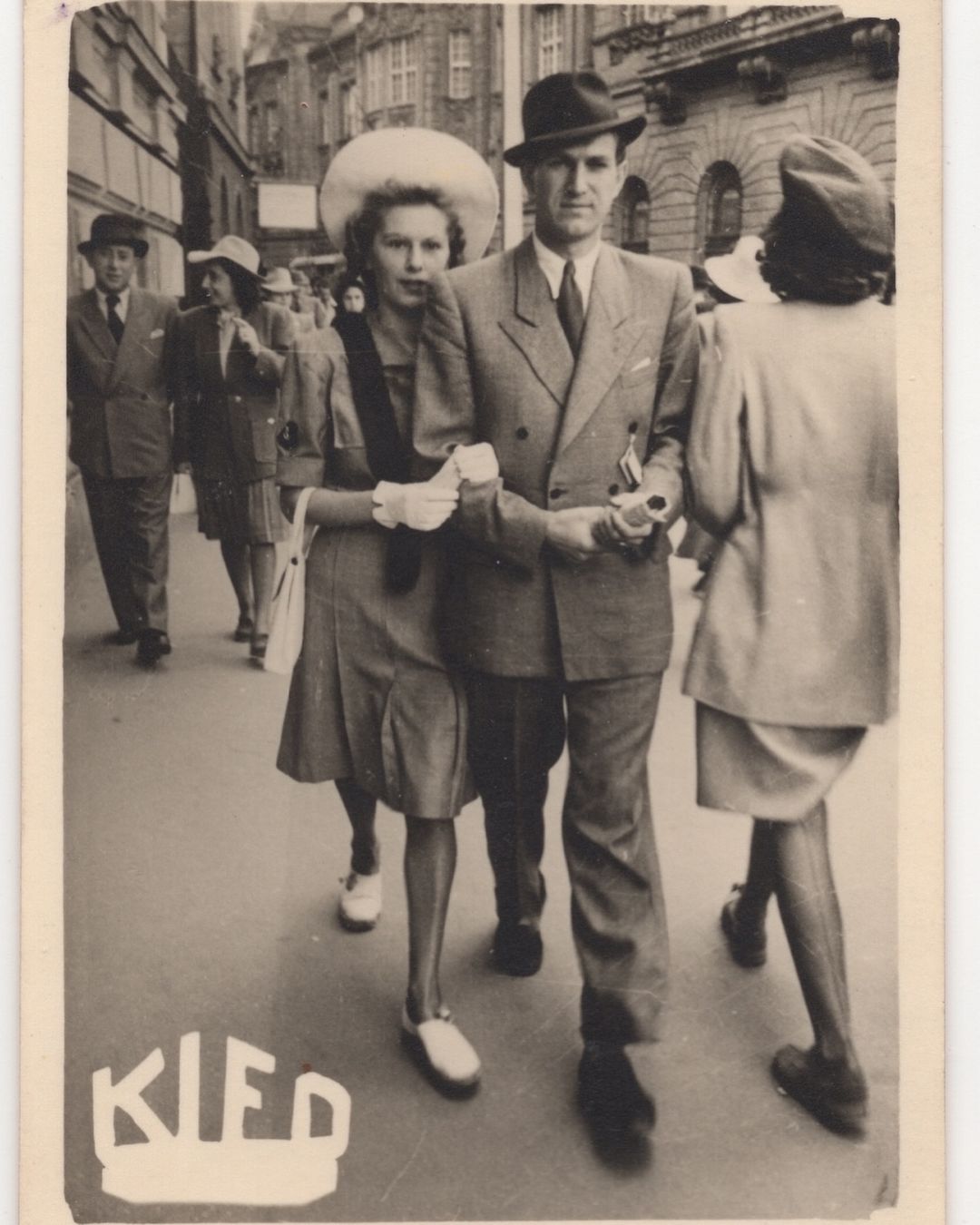 Vintage black-and-white photograph of a couple walking on a city street with other pedestrians, buildings in the background, formal attire.