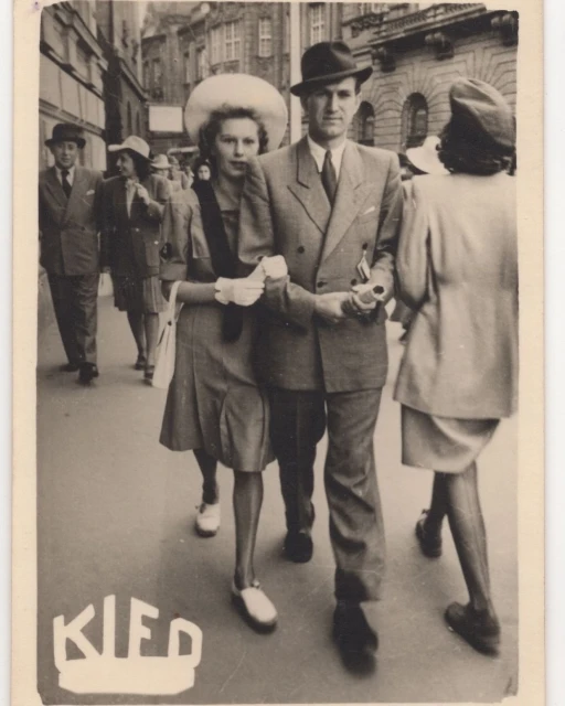 Vintage black-and-white photograph of a couple walking on a city street with other pedestrians, buildings in the background, formal attire.