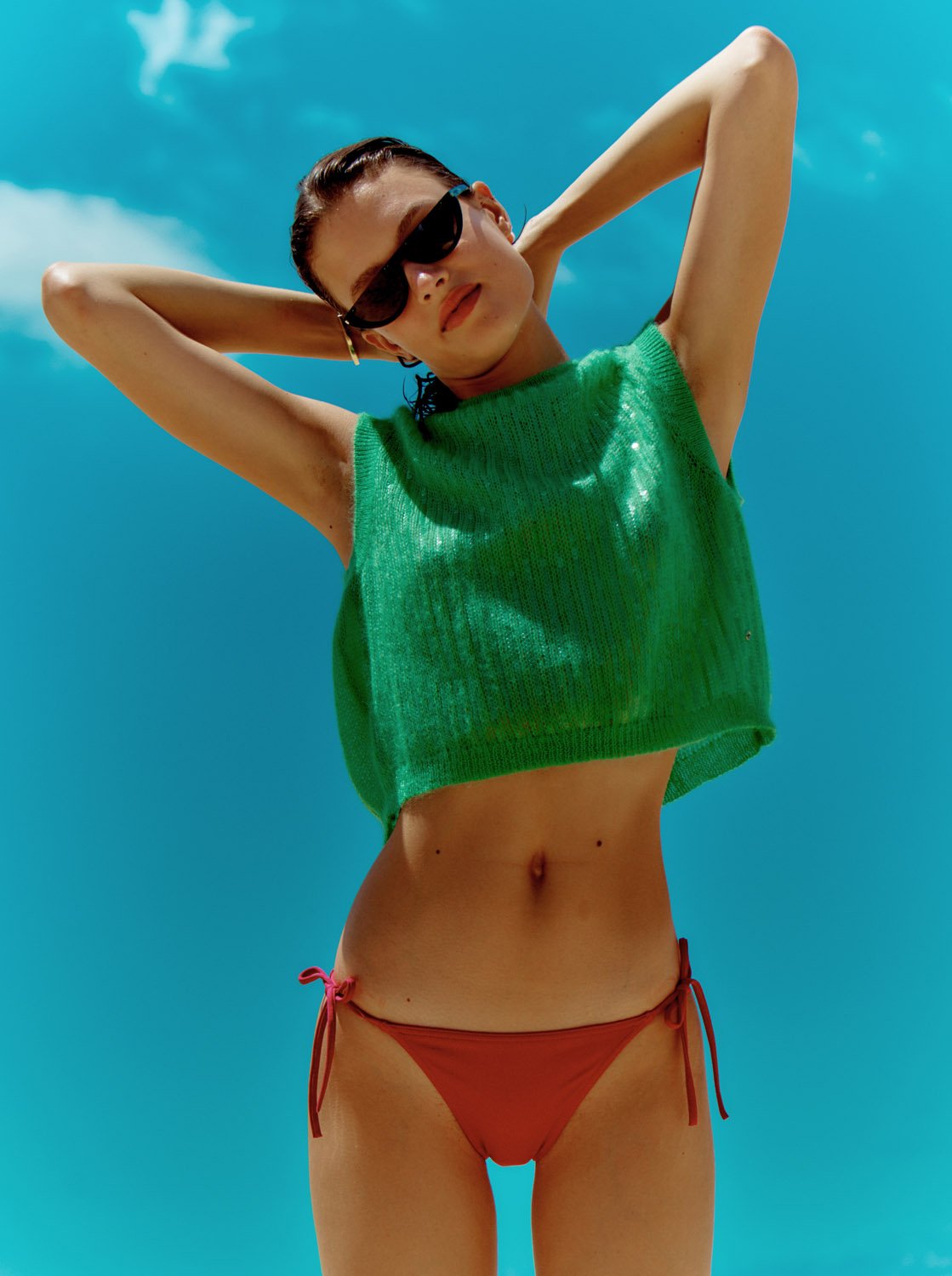 Young woman in a green knitted top and red bikini bottoms poses with hands behind her head against a bright blue sky in an eye-level shot.
