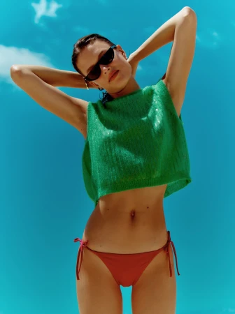 Young woman in a green knitted top and red bikini bottoms poses with hands behind her head against a bright blue sky in an eye-level shot.