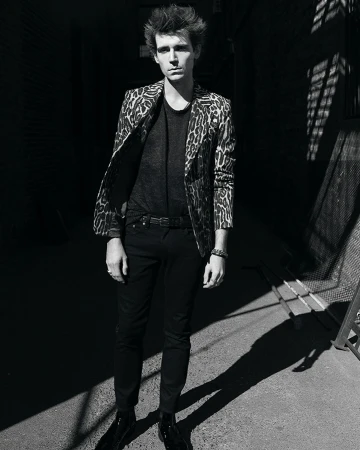 Man wearing a patterned animal print blazer and dark trousers standing outdoors in an alleyway by Adrian Mesko