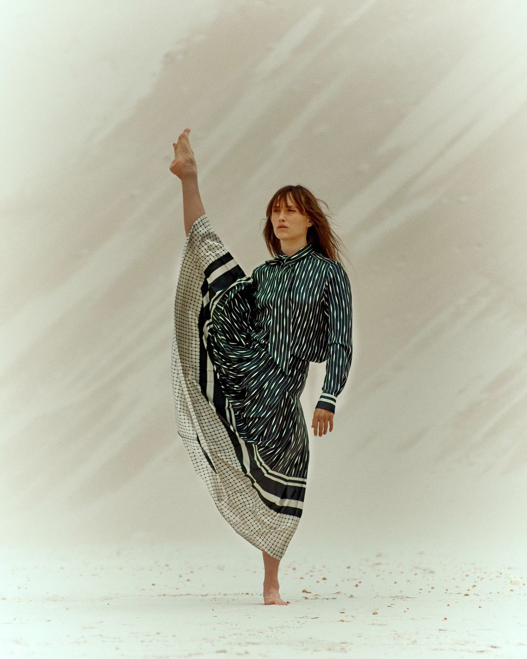 Woman in striped shirt and draped skirt strikes a barefoot pose with one leg extended upward against a light, neutral-toned backdrop.