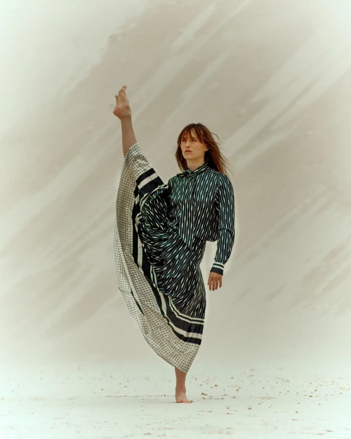 Woman in striped shirt and draped skirt strikes a barefoot pose with one leg extended upward against a light, neutral-toned backdrop.