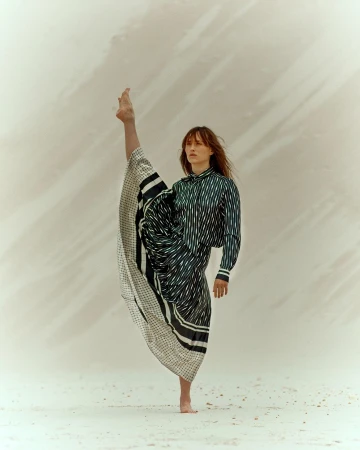 Woman in striped shirt and draped skirt strikes a barefoot pose with one leg extended upward against a light, neutral-toned backdrop.