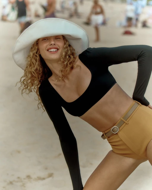 A blonde woman in a long-sleeved black crop top and yellow shorts, wearing a white hat, smiles on a sandy beach with blurred people in background.