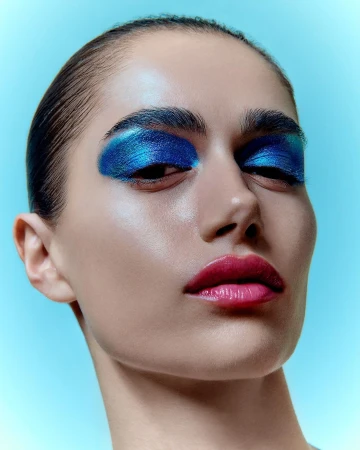 Close-up portrait of a woman with vibrant blue eyeshadow and glossy pink lips against a bright blue background by Adrian Mesko.