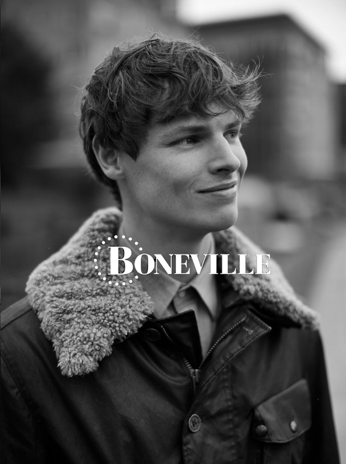 Monochrome portrait of a handsome young adult male in a jacket with a fur collar, looking off-camera, with a blurred building background.