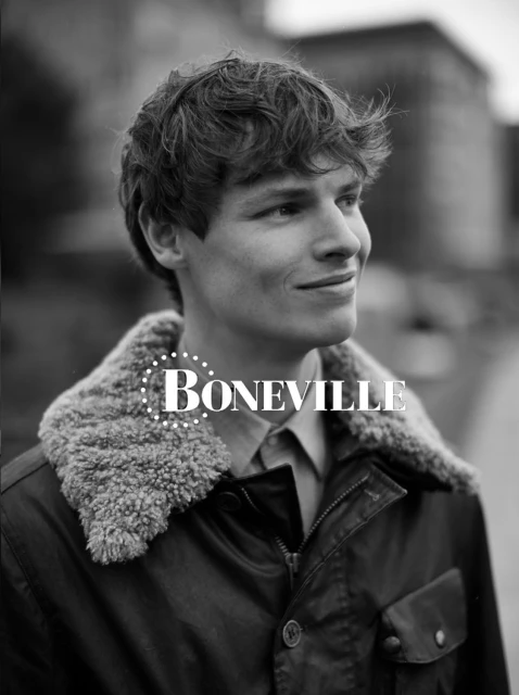 Monochrome portrait of a handsome young adult male in a jacket with a fur collar, looking off-camera, with a blurred building background.