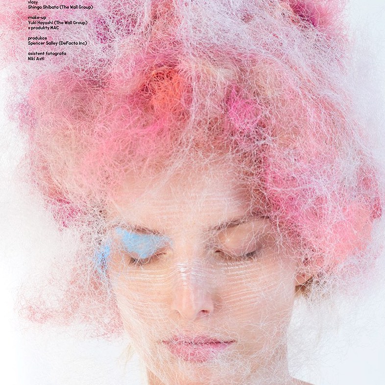 Artistic close-up of a woman with her eyes closed, adorned with strands of pink and white decorative material on her head and face.