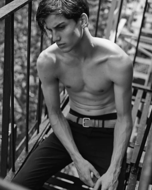 Black and white portrait of shirtless male model sitting on metal stairs outdoors, showcasing toned physique, clean style and neutral expression.