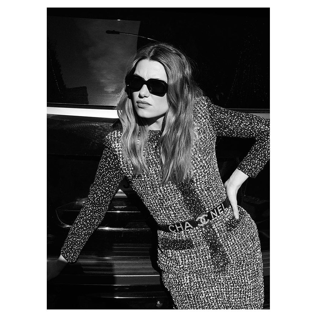 A black and white close-up of a young woman wearing sunglasses and a patterned dress with a Chanel belt, leaning against a car.