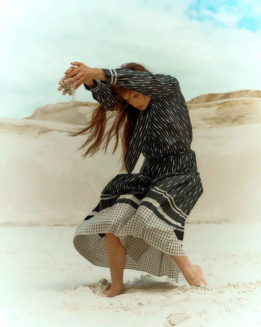 A young female model poses barefoot on white sands in a striped dress, arms raised as she holds a pile of sand. Cloudy blue sky above.