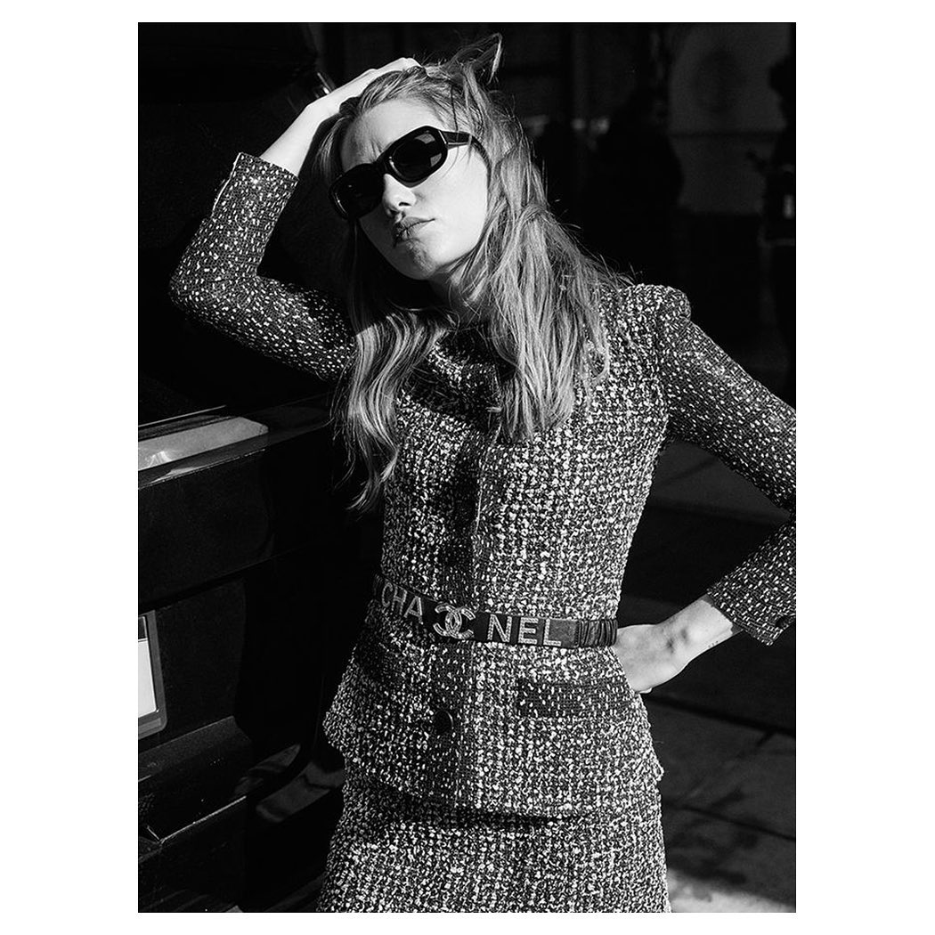 Black-and-white fashion shot of a blonde woman in a tweed suit and sunglasses leaning against a car, posing with a hand on her head.
