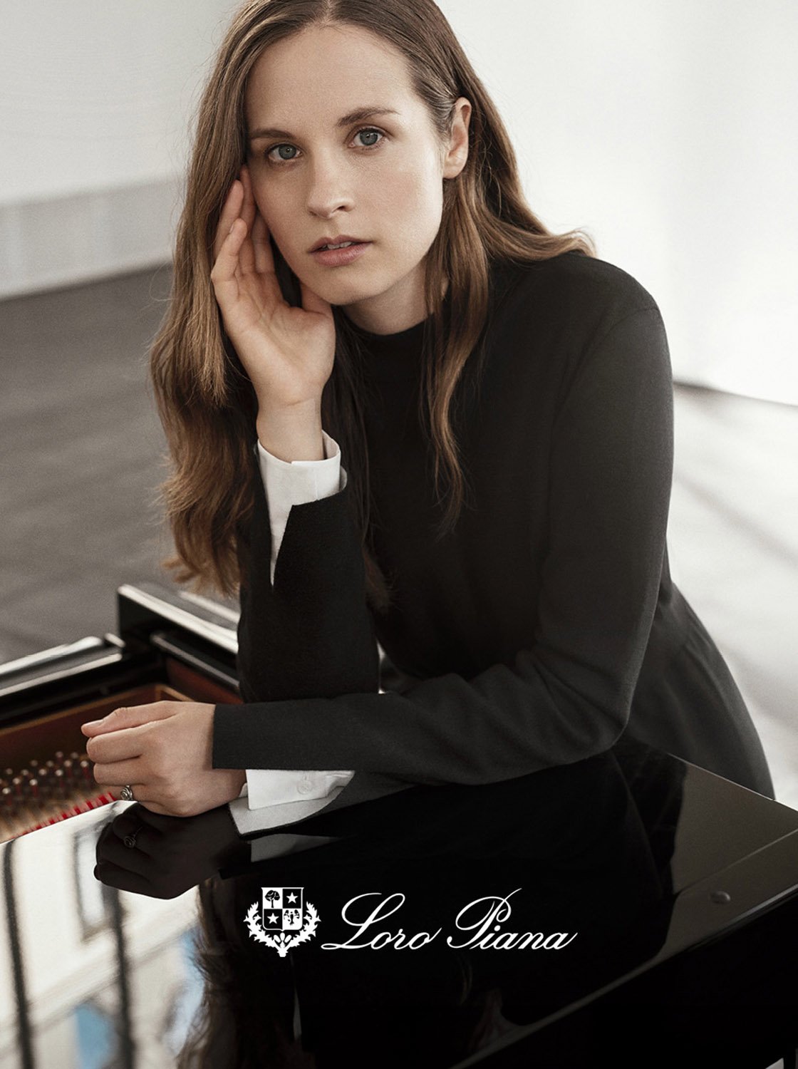 Elegant woman in black leaning on a glossy black grand piano in a bright room, featuring the Loro Piana logo at the bottom.
