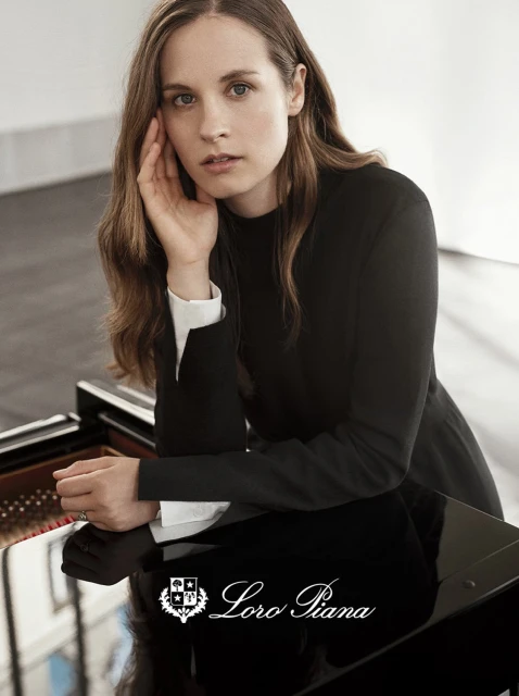 Elegant woman in black leaning on a glossy black grand piano in a bright room, featuring the Loro Piana logo at the bottom.