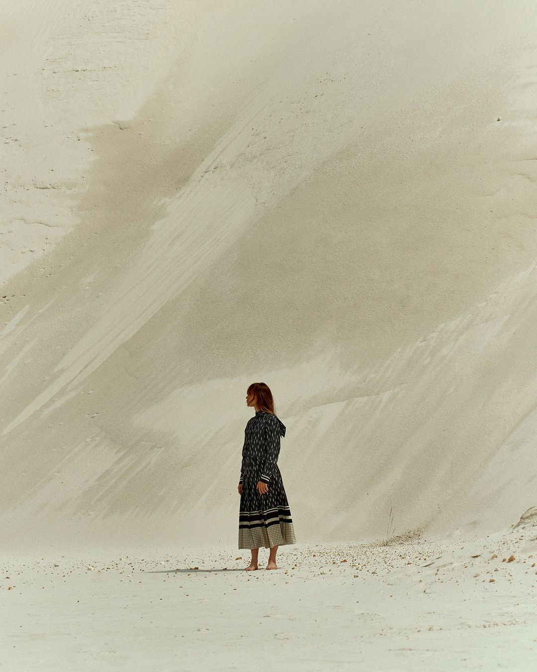 A woman stands on bare feet in a long patterned dress in the desert with large sand dunes behind her, looking at the open space.