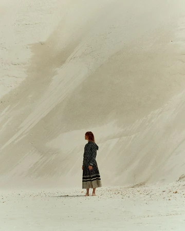 A woman stands on bare feet in a long patterned dress in the desert with large sand dunes behind her, looking at the open space.