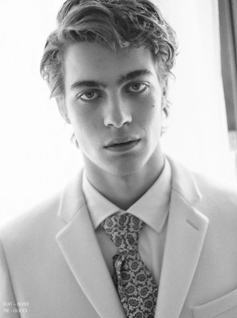 Monochrome portrait of a young adult man in a light suit and patterned tie, captured in a close-up shot with soft, neutral lighting.