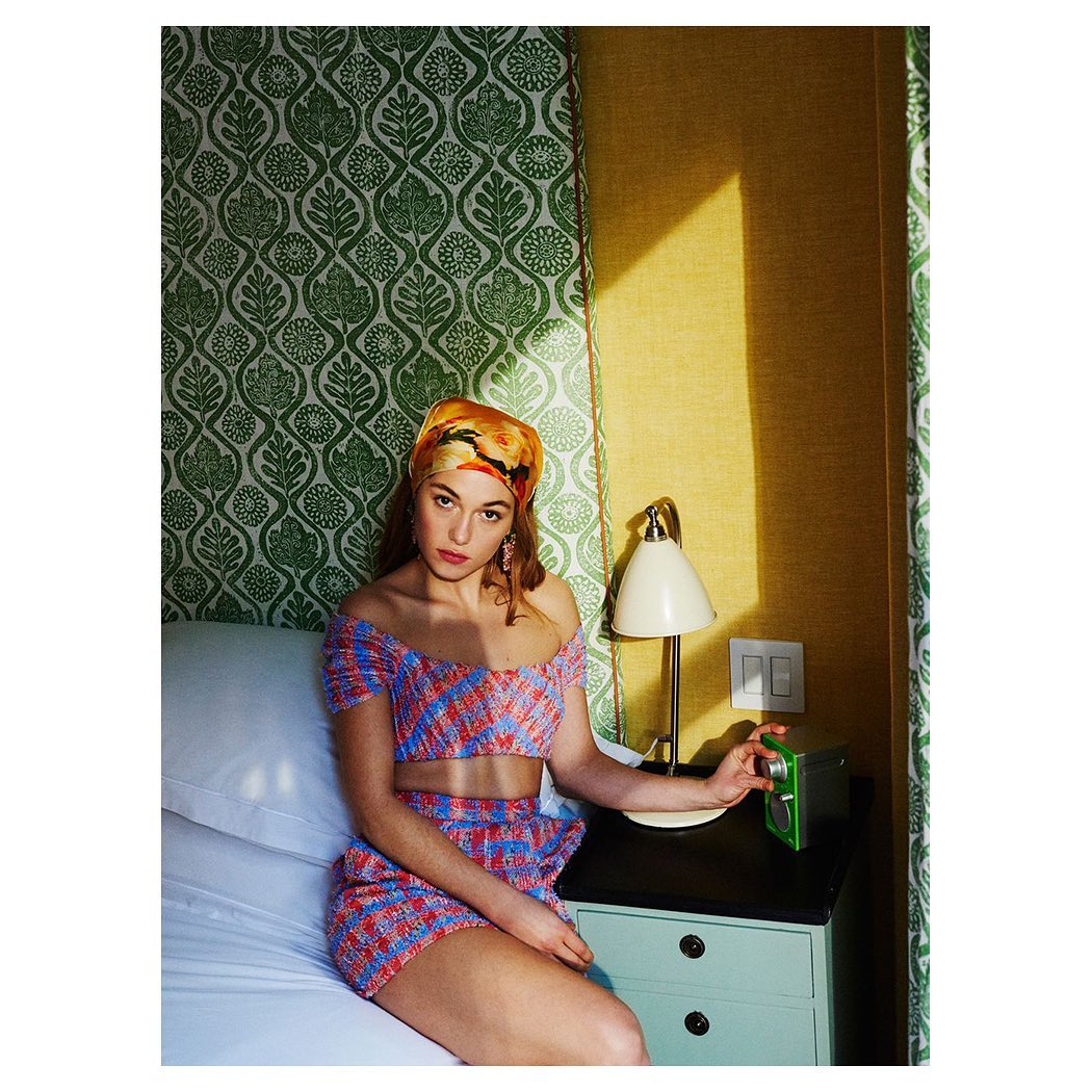 Young woman in a patterned crop top and skirt sits on a bed in a hotel room with green wallpaper and turns the dial on an alarm clock.