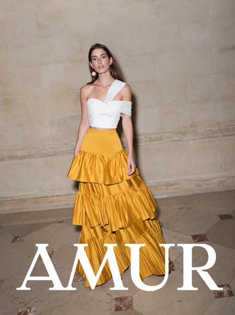 Adult female model poses in a bright photo studio, wearing a white crop top and a gold tiered, ruffled couture gown by AMUR.