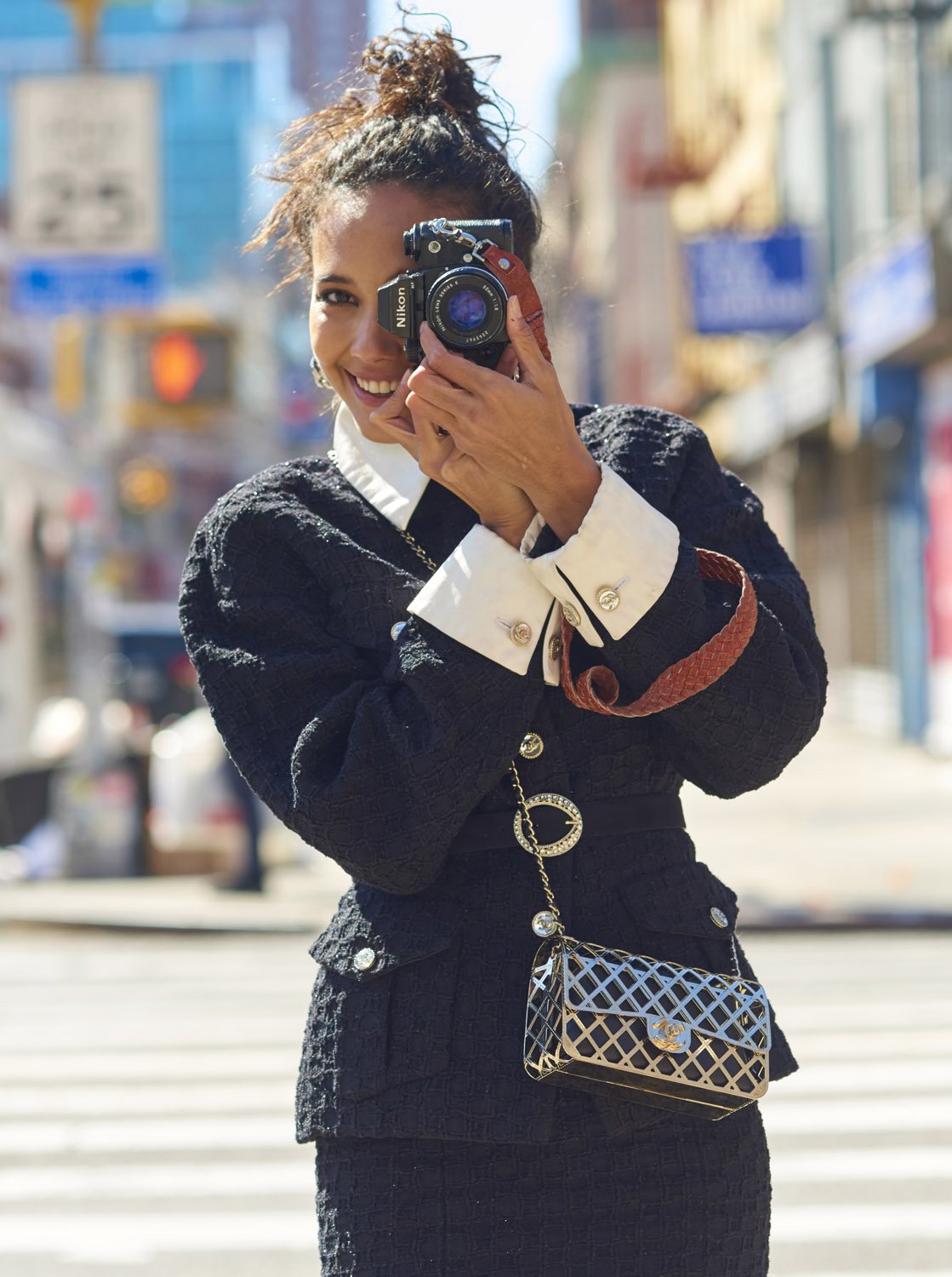 A happy woman in a tweed suit smiles at the camera while holding a vintage Nikon camera in front of her face on a city street.